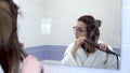 A young woman in glasses straightens her hair with a curling iron in front of a mirror. A girl in a white coat makes a Royalty Free Stock Photo