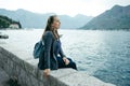 Young woman in gray cardigan and blue backpack sits near the sea Royalty Free Stock Photo