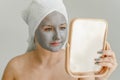 Young woman with gray clay cosmetic mask on her face looks in mirror, her hair and body wrapped in towel. Royalty Free Stock Photo