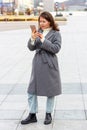 Young woman in gray coat writes message in smartphone, background of the city Royalty Free Stock Photo