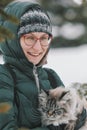 Young woman in green down jacket and glasses holding a large cat in the winter forest