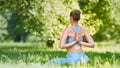 Young woman with hair bun in blue tracksuit practices yoga pose joining hands on back on green lawn