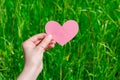 Young woman hands hold empty heart shaped pink paper piece with copy space on green grass background