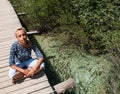 A young woman has a rest, sitting with her legs crossed, on the wooden bridge over a clear river. Royalty Free Stock Photo