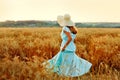 A young woman in a hat and a blue dress runs in a wheat field. The lady enjoys the sunset and nature