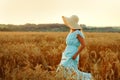 A young woman in a hat and a blue dress runs in a wheat field. The lady enjoys the sunset and nature