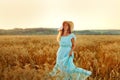 A young woman in a hat and a blue dress is spinning in a wheat field. The lady enjoys the sunset and nature