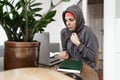 Young woman with hat and hoodie in cold home working with laptop. Energy crisis in winter