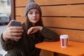 Young woman in hat and sweater sitting on the outdoor cafe, using smartphone, making selfie and drinking coffee Royalty Free Stock Photo