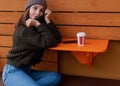 Young woman in hat and sweater sitting on the outdoor cafe, using smartphone, making selfie and drinking coffee Royalty Free Stock Photo