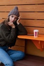 Young woman in hat and sweater sitting on the outdoor cafe, using smartphone, making selfie and drinking coffee Royalty Free Stock Photo