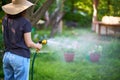 Young woman in hat watering flowers and plants in garden with hose in sunny blooming backyard Royalty Free Stock Photo