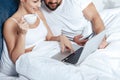 Young woman having coffee and using laptop with boyfriend in bed Royalty Free Stock Photo