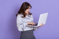 Young woman having trouble with laptop, posing against lilac wall and holding notebook with shocked and astonished facial Royalty Free Stock Photo