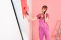 young woman in headband and working overall talking on smartphone and painting wall in red by paint Royalty Free Stock Photo