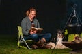 Young woman and her Australian Shepherd outside by a campfire. Reading a book at dusk. Bread, cheese and wine on the Royalty Free Stock Photo