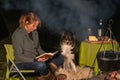 Young woman and her Australian Shepherd outside by a campfire. Reading a book at dusk. Bread, cheese and wine on the Royalty Free Stock Photo