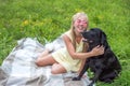 Young woman with her big black dog is sitting on a blanket on the grass in an embrace. Portrait of blonde woman in pink glasses