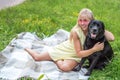 Young woman with her big black dog is sitting on a blanket on the grass in an embrace. Portrait of blonde woman in pink glasses