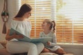 Young woman and her daughter reading book near window at home Royalty Free Stock Photo