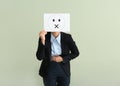 Young woman hiding face behind sheet of paper with drawn emoticon on light background Royalty Free Stock Photo