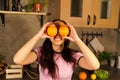 Young woman hiding her face behind fruits in kitchen. Adult lady flirting and looking out of oranges at camera. Royalty Free Stock Photo