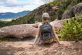 Young woman in hiking clothes, resting on stones near natural water pool, blurred trees and mountains background, view form behind Royalty Free Stock Photo