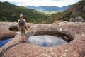 Young woman in hiking clothes, standing on stones near natural water pool, blurred trees and mountains background, view form Royalty Free Stock Photo