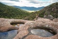 Young woman in hiking clothes, standing on stones near natural water pool, blurred trees and mountains background, view form Royalty Free Stock Photo