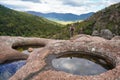 Young woman in hiking clothes, standing on stones near natural water pool, blurred trees and mountains background, view form Royalty Free Stock Photo