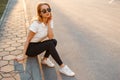 Young woman hipster in stylish sunglasses in a white polo shirt in black jeans in white fashionable sneakers sits on the asphalt Royalty Free Stock Photo