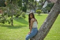 Young woman, Hispanic, beautiful, brunette, with daisy t-shirt and jeans, leaning on a tree trunk, calm and relaxed. Concept of Royalty Free Stock Photo