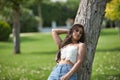 Young woman, Hispanic, beautiful, brunette, with daisy t-shirt and jeans, leaning on a tree trunk, calm and relaxed. Concept of Royalty Free Stock Photo