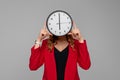 Young woman Holding Big Clock Covering her Face, she wonders how much time passed, standing in red blazer against gray background Royalty Free Stock Photo