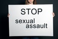 Young woman holding card with words STOP SEXUAL ASSAULT against dark background