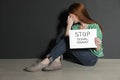 Young woman holding card with words STOP SEXUAL ASSAULT while sitting near wall. Space for text