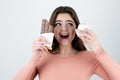Young woman holding chocolate bar in one hand and cup cake with cream in another looking surprised standing on isolated white Royalty Free Stock Photo