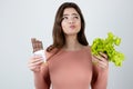 Young woman holding chocolate bar in one hand and green salad in another looking hesitant standing on isolated white background, Royalty Free Stock Photo