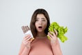 Young woman holding chocolate bar in one hand and green salad in another looking surprised standing on isolated white background, Royalty Free Stock Photo