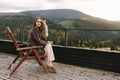 A young woman holding coffee cup while sittinhg on chair on balcony , looking at mountains and green nature Royalty Free Stock Photo