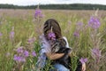 Young woman holding gray fluffy senior dog in hands on meadow with flowers of fireweed or great willowherb, pet love Royalty Free Stock Photo