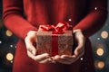 Young woman holding in hands a Christmas gift in shiny glittering package with red ribbon. Festive blurred background with golden