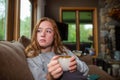 Young woman holding a hot cup of tea and looking out the window with a concerned look on her face