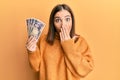 Young woman holding 5000 japanese yen banknotes covering mouth with hand, shocked and afraid for mistake