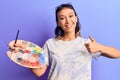 Young woman holding paintbrush and palette smiling happy and positive, thumb up doing excellent and approval sign Royalty Free Stock Photo