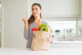 Young woman holding paper bag full of groceries pointing and showing with thumb up to the side with happy face smiling Royalty Free Stock Photo