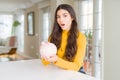 Young woman holding piggy bank scared in shock with a surprise face, afraid and excited with fear expression Royalty Free Stock Photo