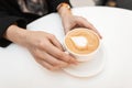 Young woman holds in hands a cup with a hot sweet cappuccino sitting at a white table in a cafe. Great morning for coffee gourmets Royalty Free Stock Photo