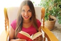 Young woman at home sitting on comfortable chair relaxing in her living room reading book. Looks at camera Royalty Free Stock Photo