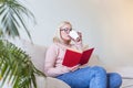 Young woman at home sitting on modern sofa relaxing in her living room reading book and drinking coffee or tea. White cozy bed and Royalty Free Stock Photo